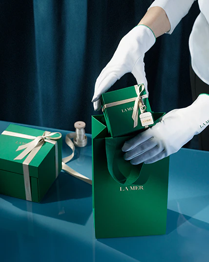 Photograph of persons hands with gloves packing the la mer gift box into a la mer green gift bag.